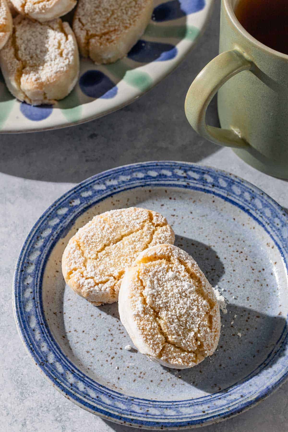 A close up of 2 baked ricciarelli cookies topped with powdered sugar on a plate in front of a mug, and the plate with the rest of the cookies.