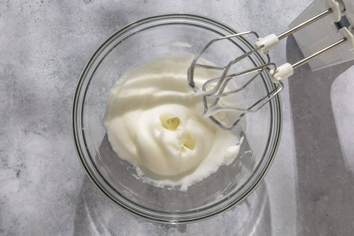 Whipped egg whites in a bowl next to a hand mixer.