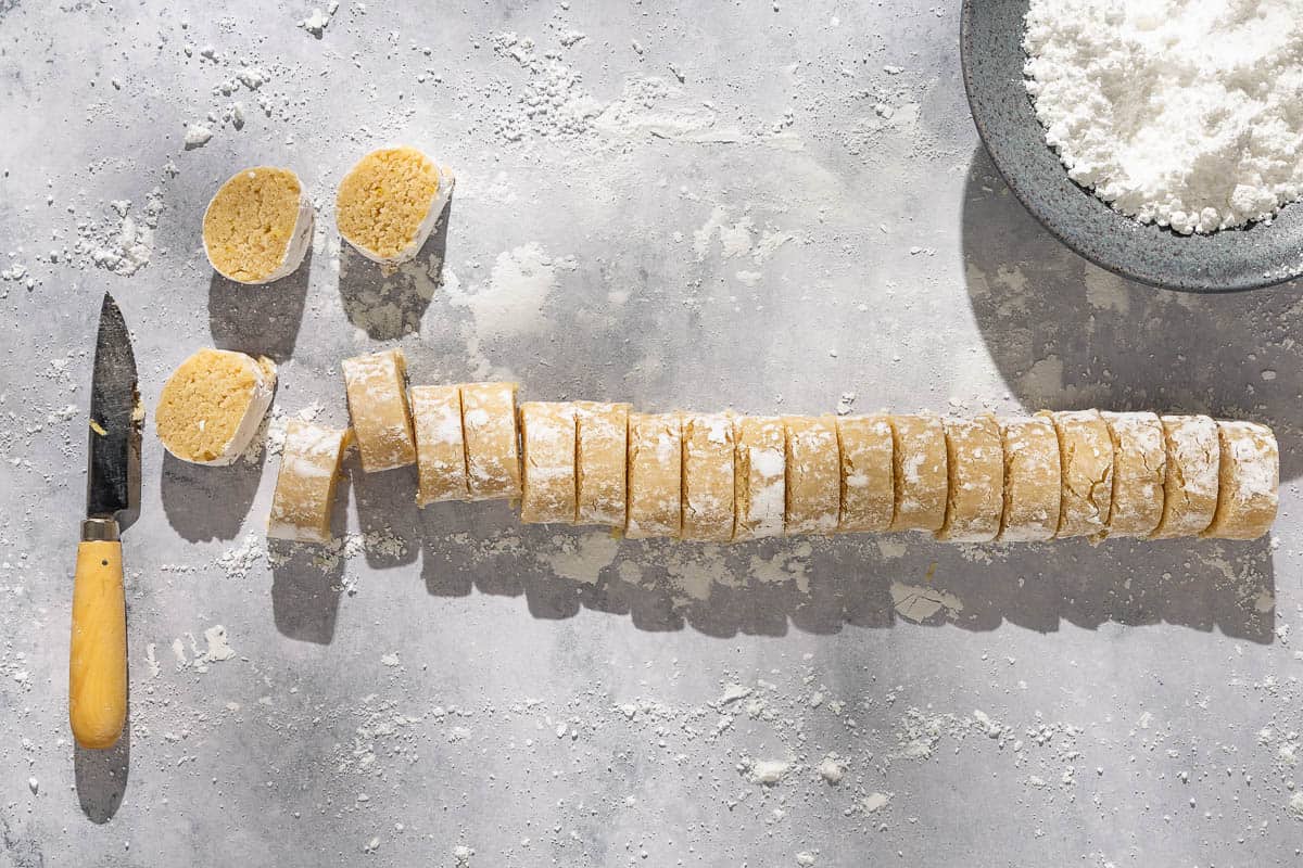 The dough for the ricciarelli cookies rolled into a log and cut into slices next to a knife and a bowl of powdered sugar.