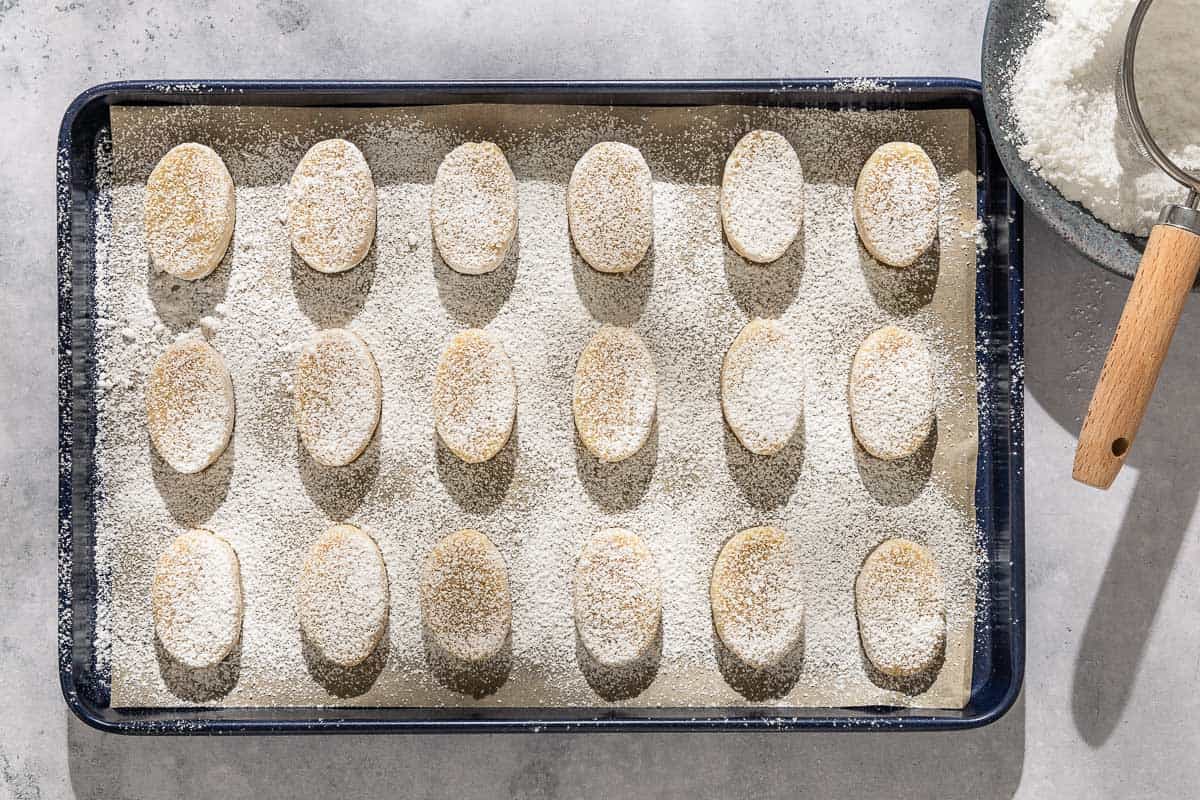 Unbaked ricciarelli cookies topped with powdered sugar on a parchment line sheet pan. Next to this is a bowl of powdered sugar with a mesh strainer.