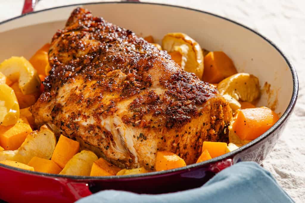 A close up of a roast pork loin with butternut squash and apples in a roasting pan.