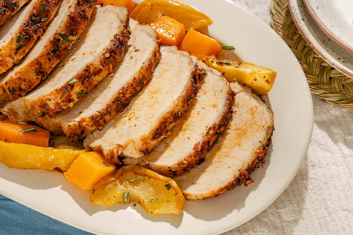 A close up of sliced roast pork loin with butternut squash and apples on a serving platter. Next to this is a stack of 2 plates.