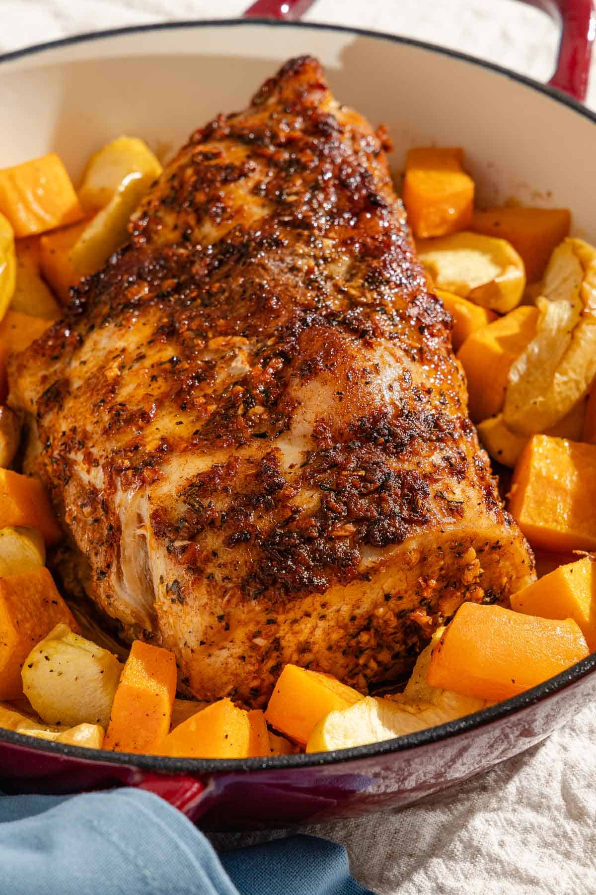 A close up of a roast pork loin with butternut squash and apples in a roasting pan.