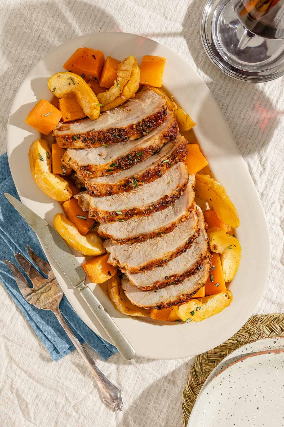 An overhead photo of sliced roast pork loin with butternut squash and apples on a serving platter with a knife. Next to this is a serving fork on a cloth napkin and a glass of wine.