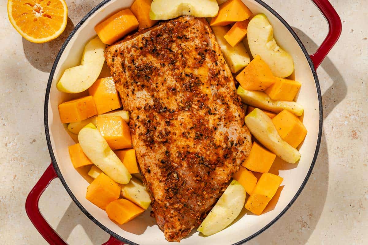 An overhead photo of a pork loin with butternut squash and apples in a roasting pan before being cooked. Next to this is an orange half.