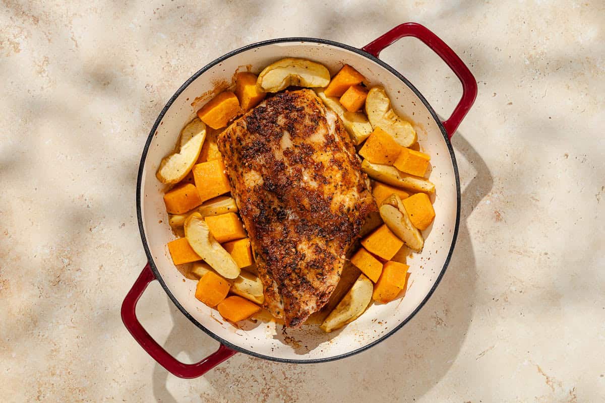 An overhead photo of a roast pork loin with butternut squash and apples in a roasting pan.