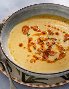 A close up of garlic soup topped with fried garlic and aleppo oil in a bowl on a plate with a spoon. Next to this is a piece of sliced bread and a small bowl of the fried garlic and aleppo oil.