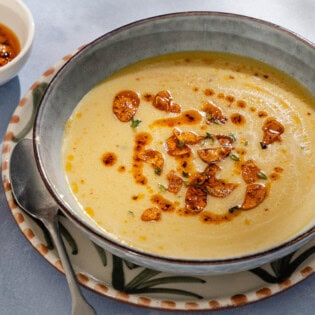 A close up of garlic soup topped with fried garlic and aleppo oil in a bowl on a plate with a spoon. Next to this is a piece of sliced bread and a small bowl of the fried garlic and aleppo oil.