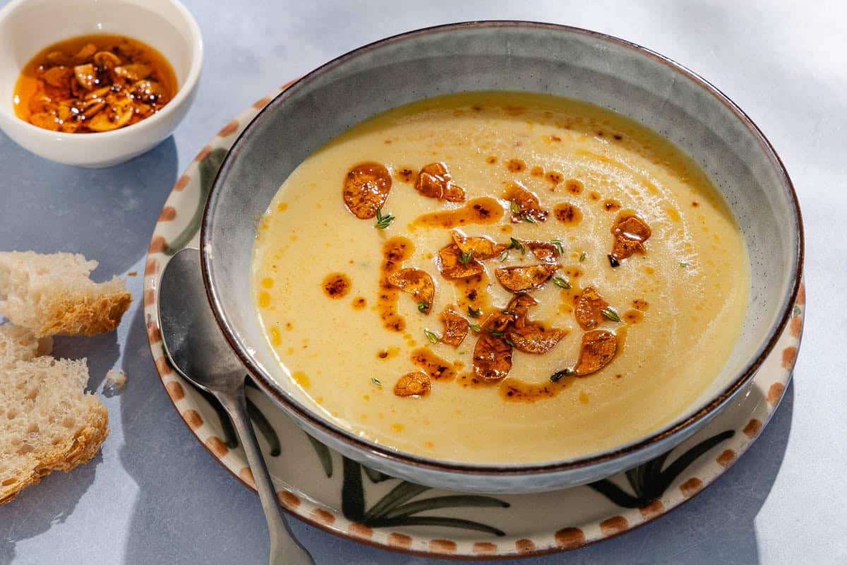 A close up of garlic soup topped with fried garlic and aleppo oil in a bowl on a plate with a spoon. Next to this is a piece of sliced bread and a small bowl of the fried garlic and aleppo oil.