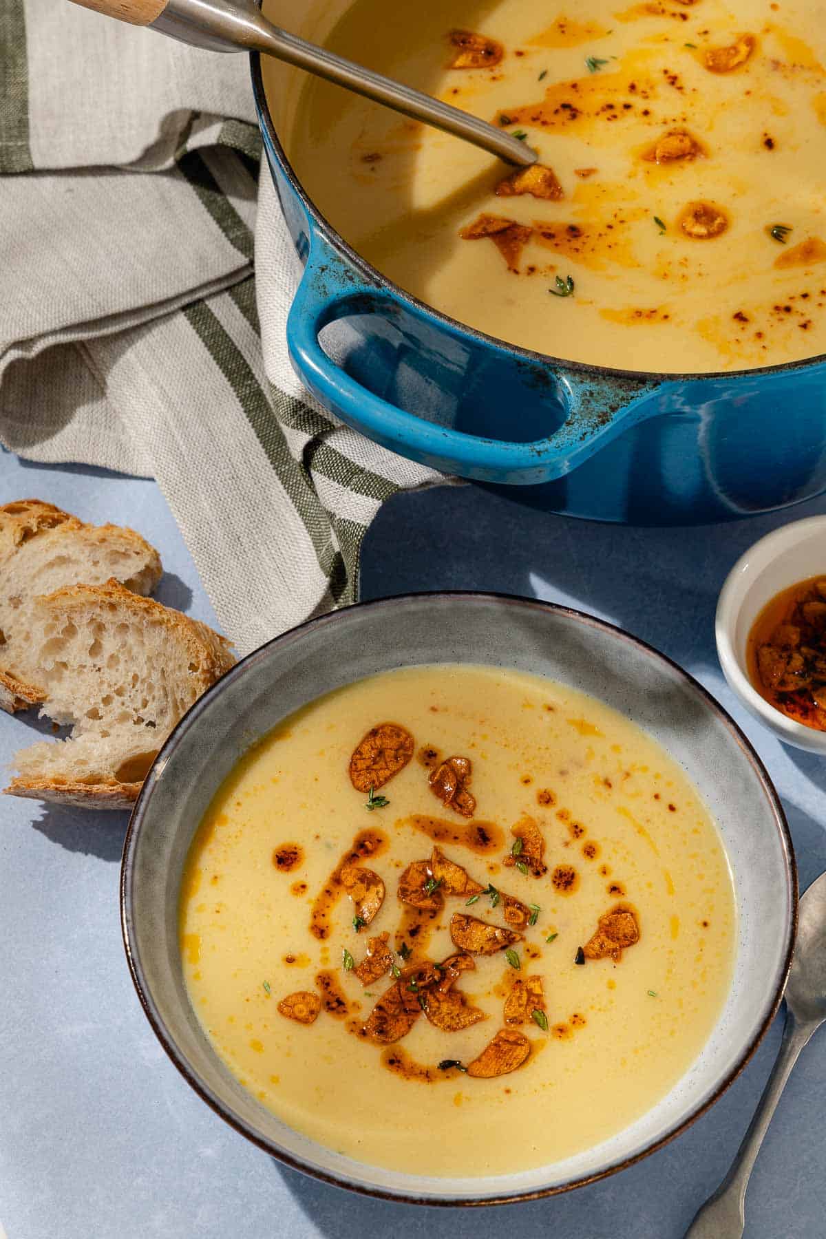 Garlic soup topped with fried garlic and aleppo oil in a bowl. Next to this is a spoon, the pot of the soup, a kitchen towel, pieces of sliced bread and a small bowl of the fried garlic and aleppo oil.