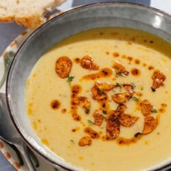 A close up of garlic soup topped with fried garlic and aleppo oil in a bowl on a plate with a spoon. Next to this is a small bowl of the fried garlic and aleppo oil