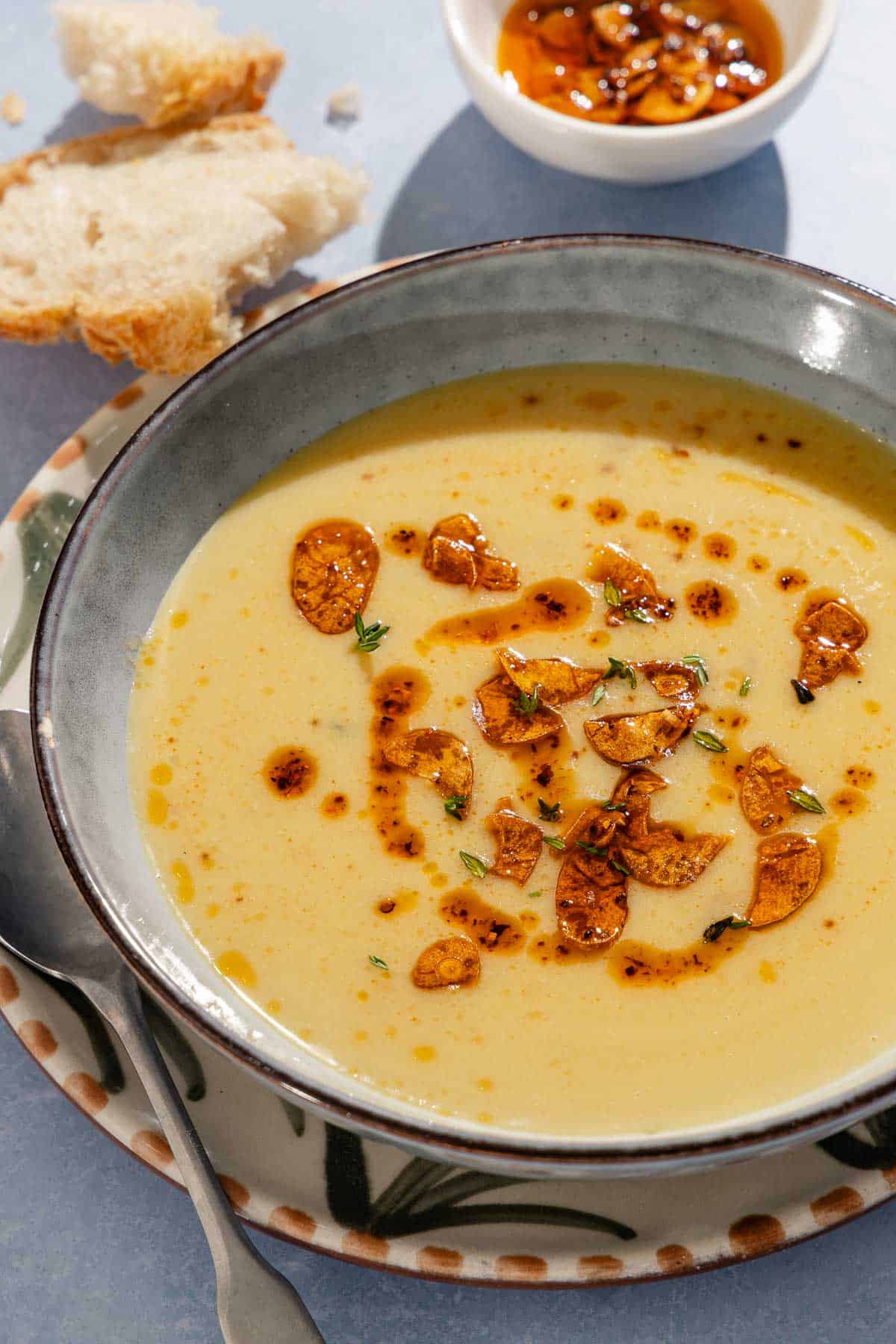 A close up of garlic soup topped with fried garlic and aleppo oil in a bowl on a plate with a spoon. Next to this is a small bowl of the fried garlic and aleppo oil