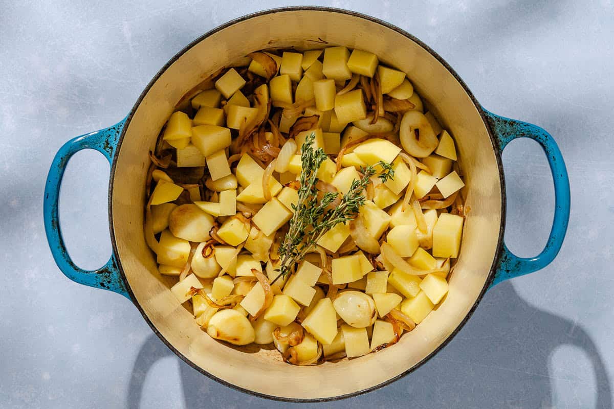 An overhead photo of caramelized onions and garlic, cubed uncooked potatoes and fresh thyme in a large pot.