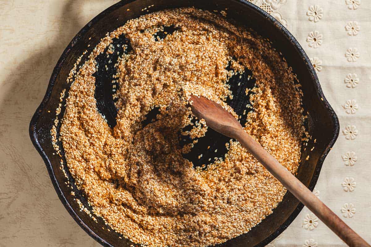 Toasted sesame seeds being mixed with honey in a cast iron skillet with a wooden spoon.