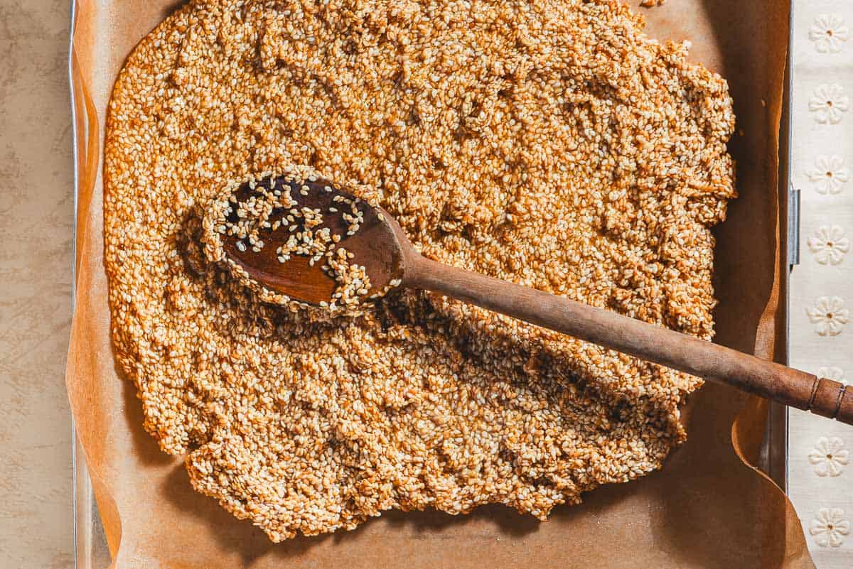 The honey and sesame seed mixture being spread evenly in a parchment-lined baking sheet with a wooden spoon.