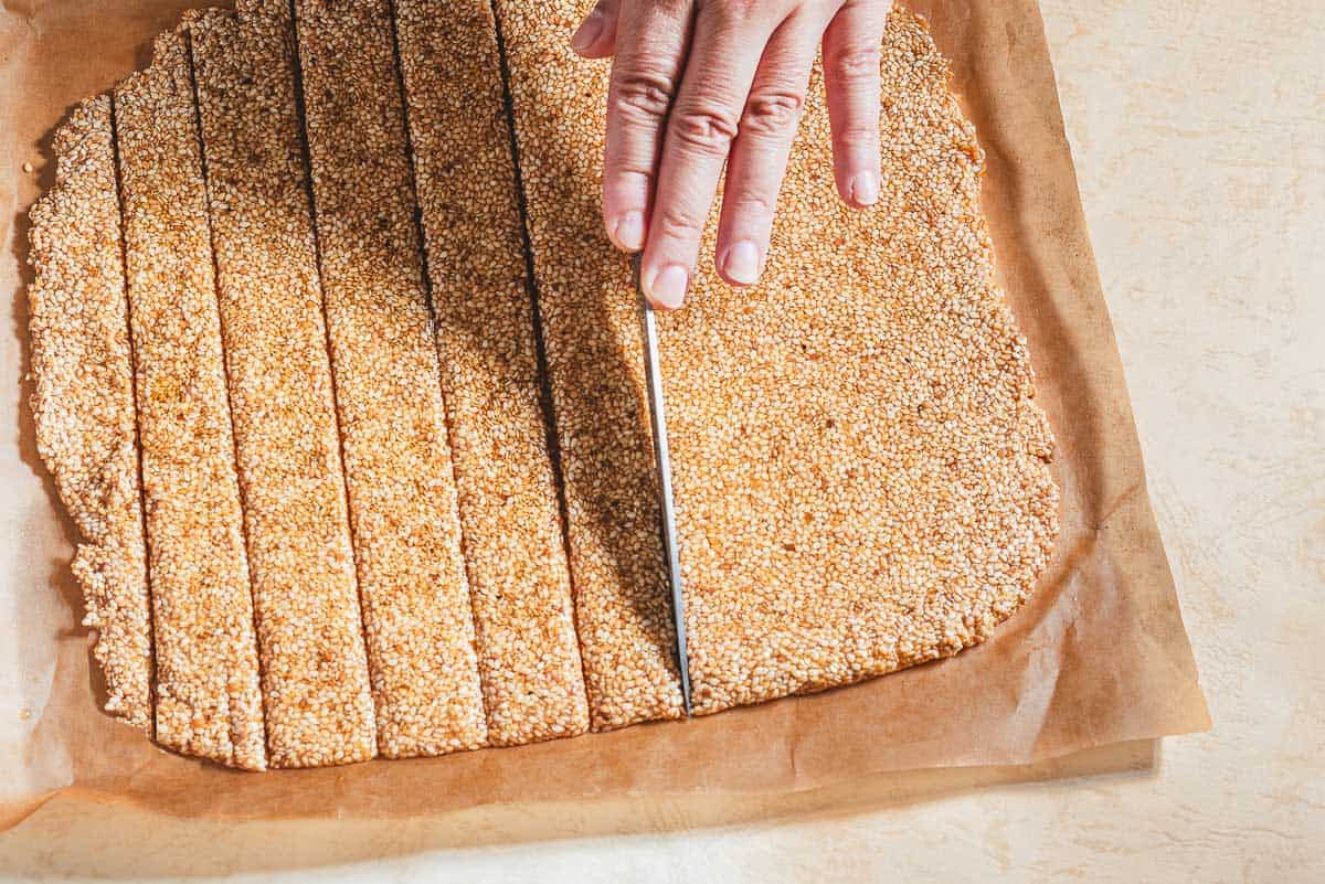 Greek sesame candy with honey an a sheet of parchment paper being sliced into individual pieces with a knife.