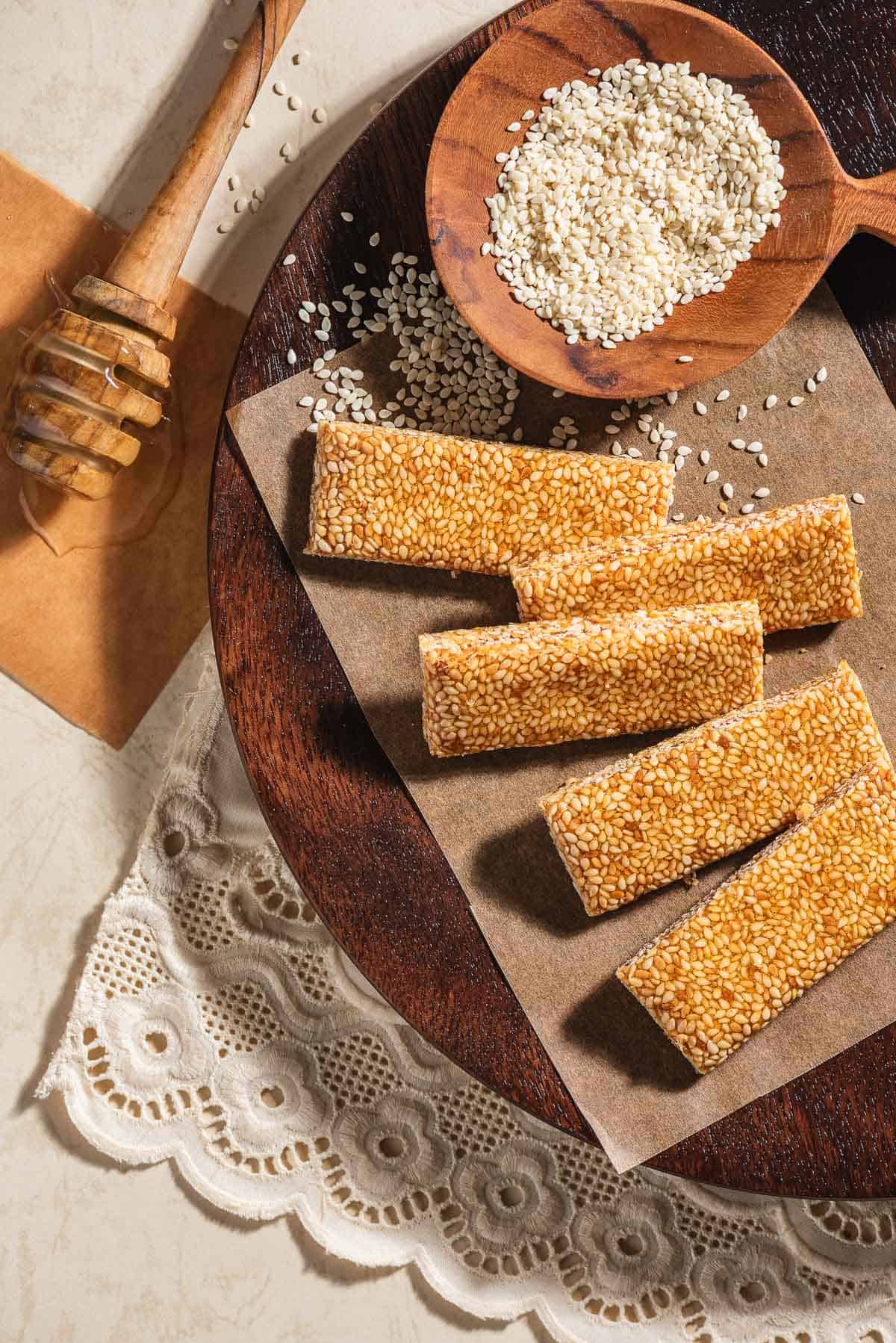 5 pieces of greek sesame candy with honey an a sheet of parchment paper with a bowl of the sesame seeds on a wooden tray. Next to this is a honey dipper.