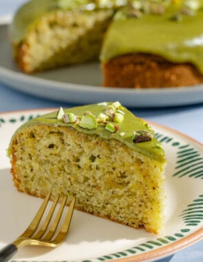 A close up of a slice of pistachio cake on a plate with a fork. In the background is the rest of the cake on a platter.