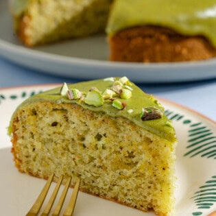 A close up of a slice of pistachio cake on a plate with a fork. In the background is the rest of the cake on a platter.