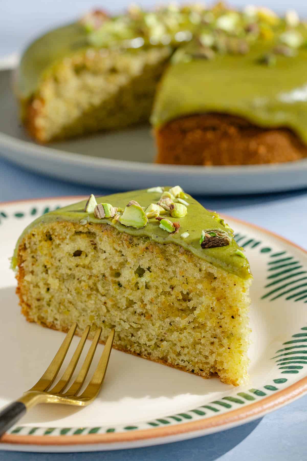 A close up of a slice of pistachio cake on a plate with a fork. In the background is the rest of the cake on a platter.