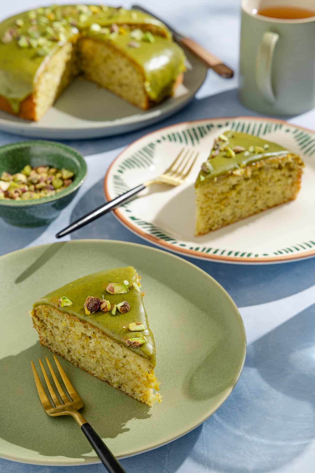 Two slices of pistachio cake on 2 plates with forks. Next to these is a bowl of pistachios, a mug of tea, and the rest of the cake on a platter with a knife.