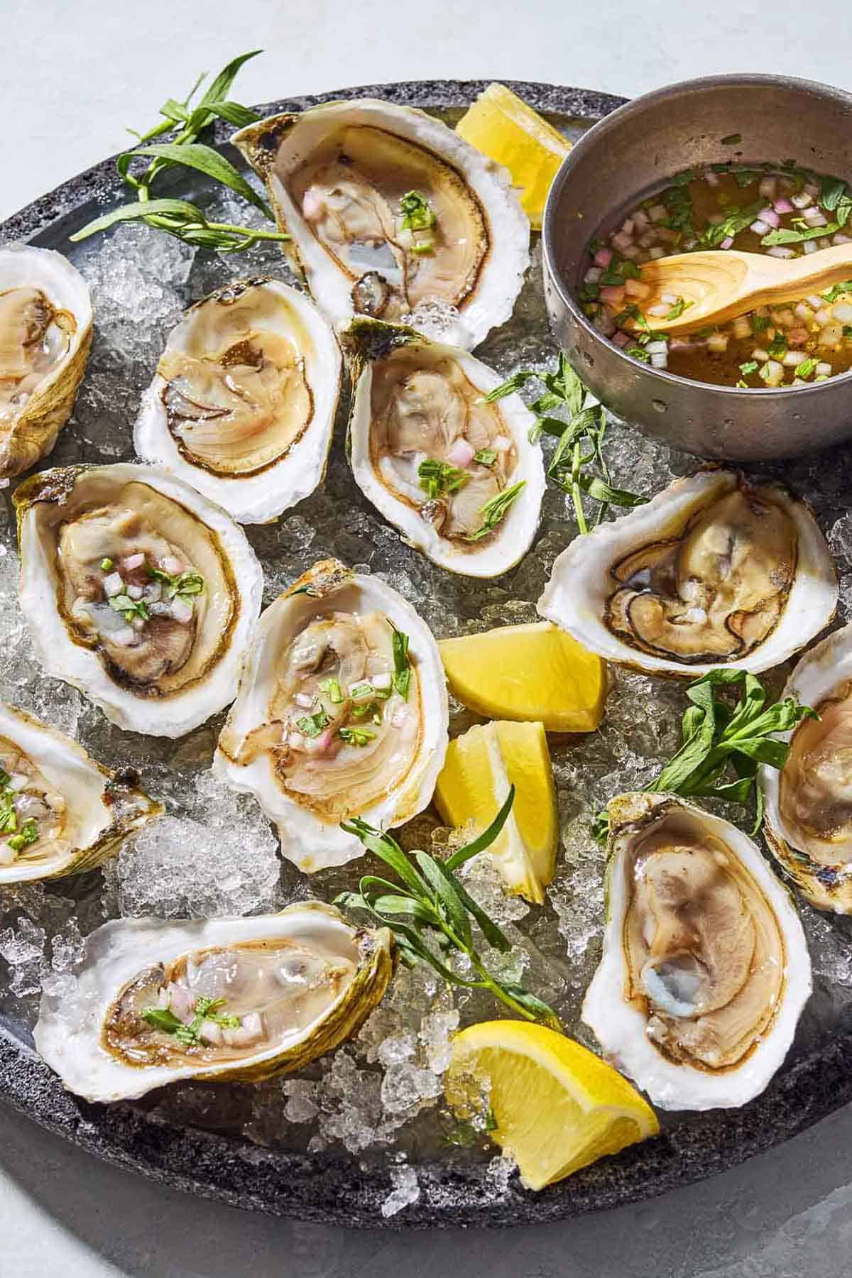 Several raw oysters with mignonette sauce on a bed of ice on a tray with lemon wedges and a bowl with the rest of the sauce and a spoon.