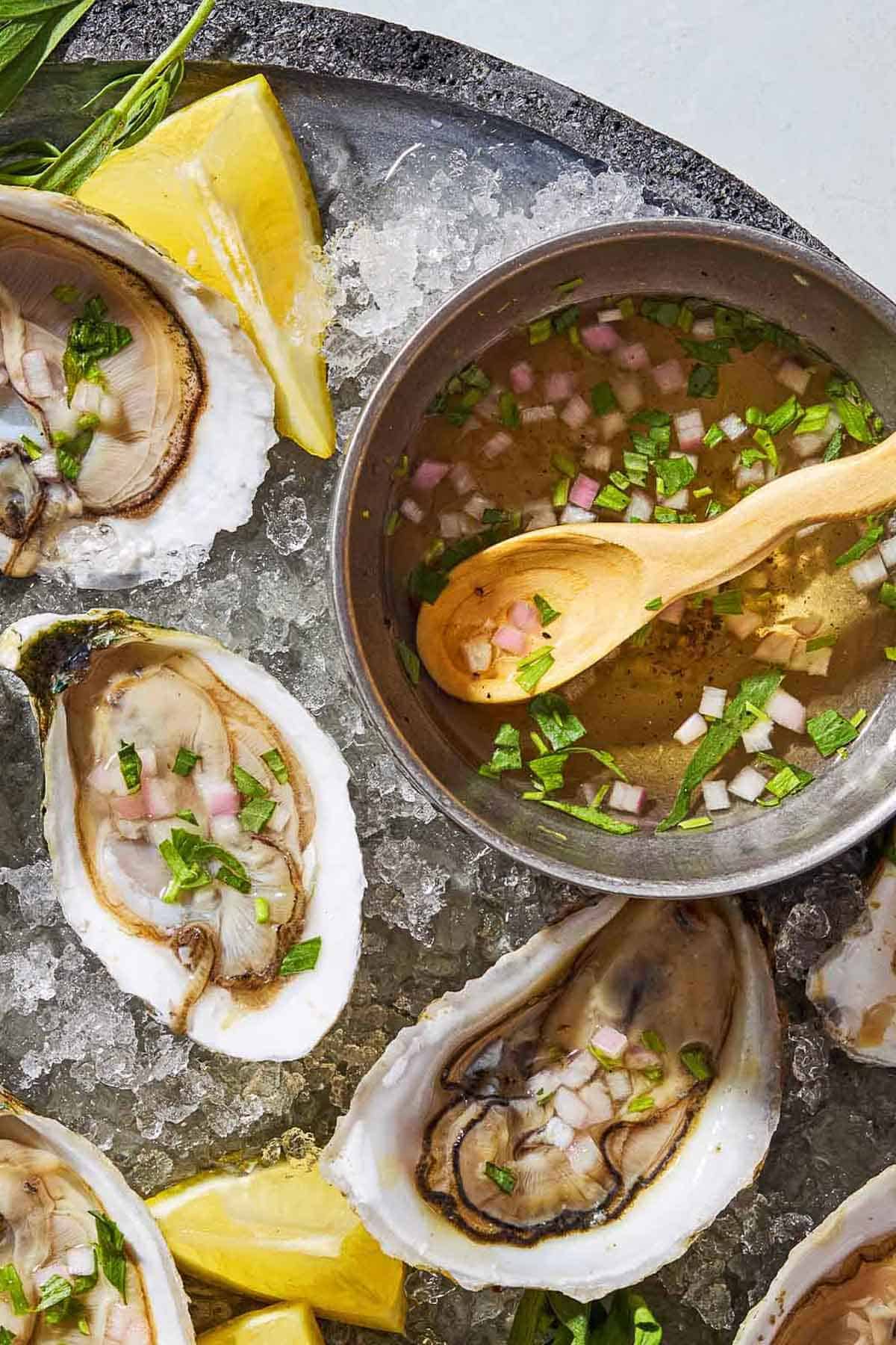 Several raw oysters with mignonette sauce on a bed of ice on a tray with lemon wedges and a bowl with the rest of the sauce and a spoon.