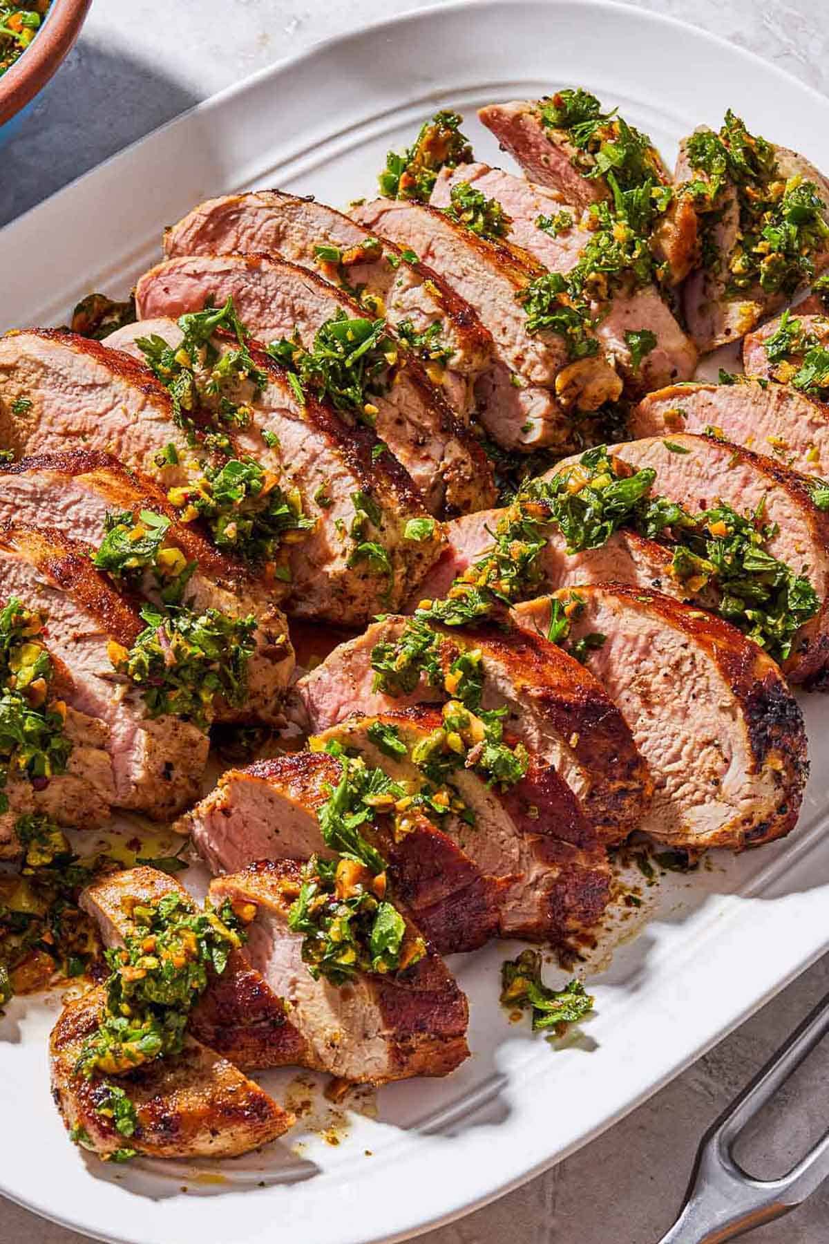 Sliced baked pork tenderloin topped with herb and pistachio salsa on a platter.