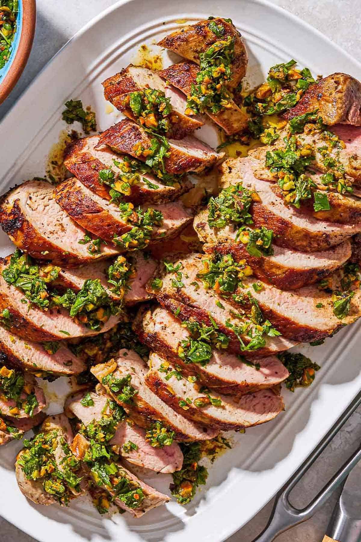 Sliced baked pork tenderloin topped with herb and pistachio salsa on a platter.