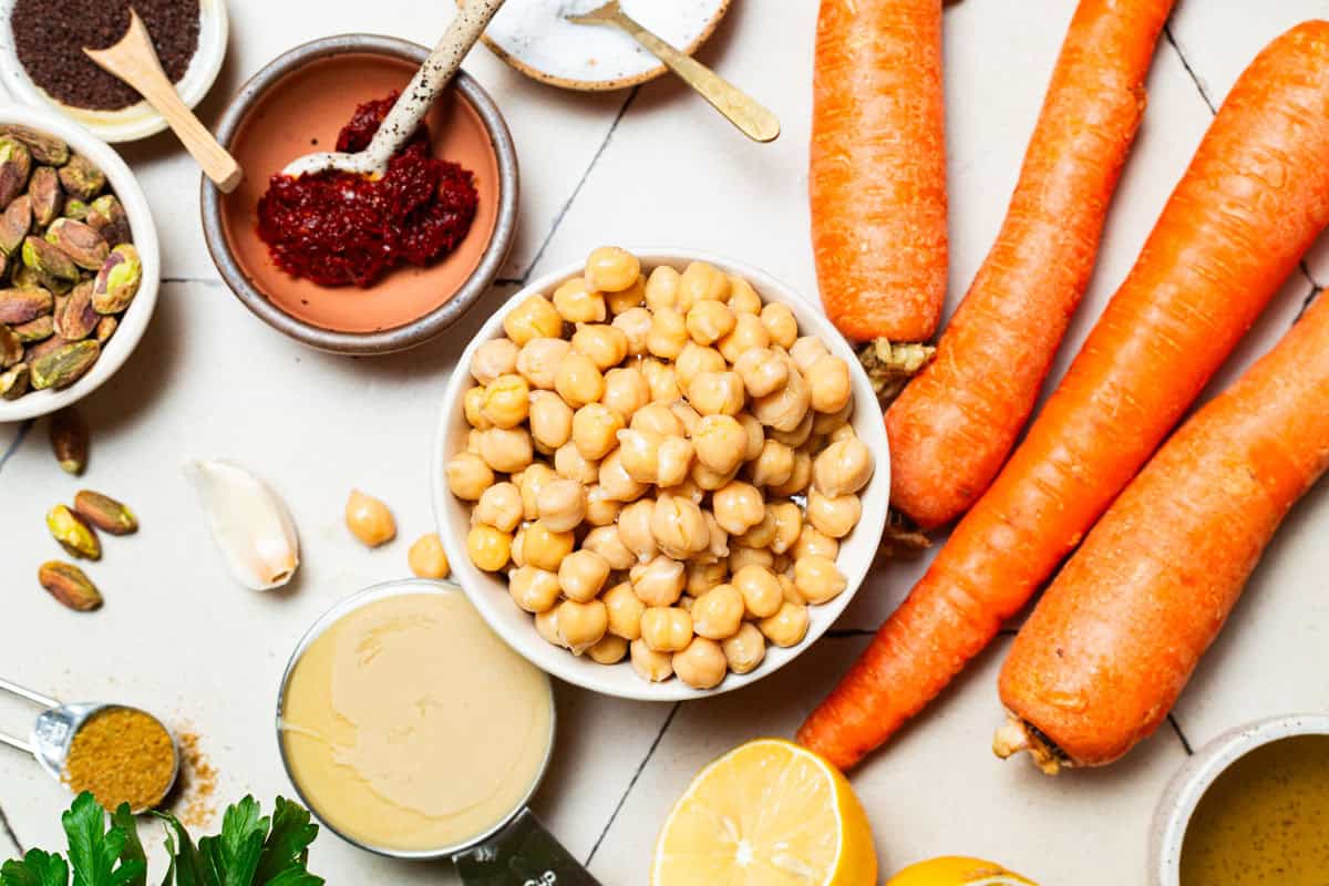 Ingredients for roasted carrot hummus including carrots, chickpeas, olive oil, salt, tahini paste, garlic, sumac, cumin, harissa paste, lemon, parsley and pistachios.