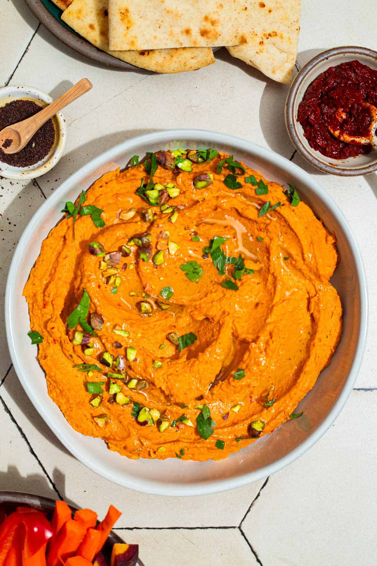 An overhead photo of roasted carrot hummus topped with chopped pistachios and parsley in a bowl. Next to this is a som pita bread and bowls of harissa paste, sumac and carrot sticks.