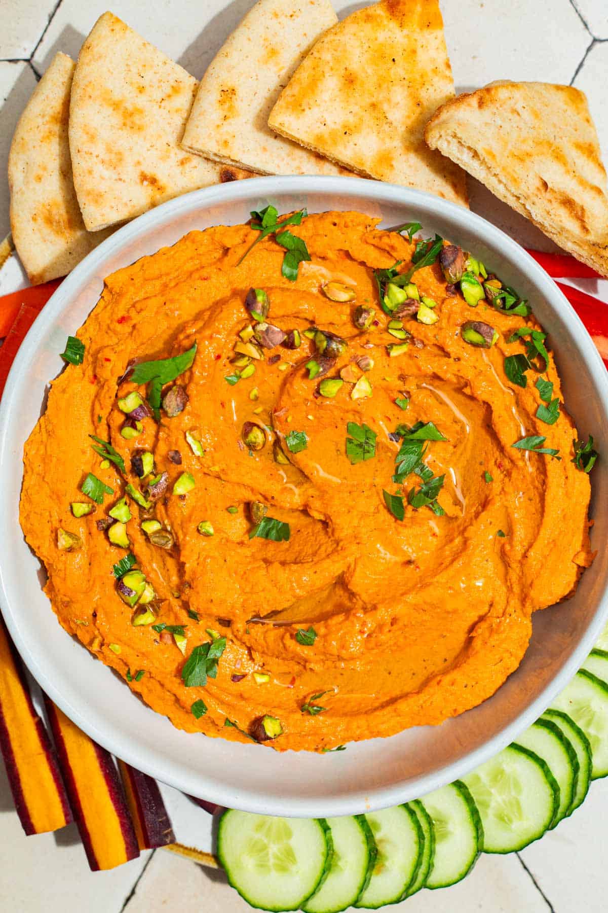 An overhead photo of roasted carrot hummus topped with chopped pistachios and parsley in a bowl on a platter surrounded by pita bread triangles, carrot sticks and cucumber slices.