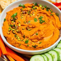 A close up of roasted carrot hummus topped with chopped pistachios and parsley in a bowl on a platter surrounded by pita bread triangles, carrot sticks and cucumber slices.