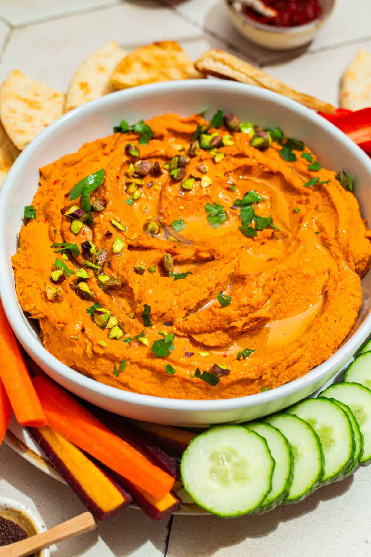 A close up of roasted carrot hummus topped with chopped pistachios and parsley in a bowl on a platter surrounded by pita bread triangles, carrot sticks and cucumber slices.