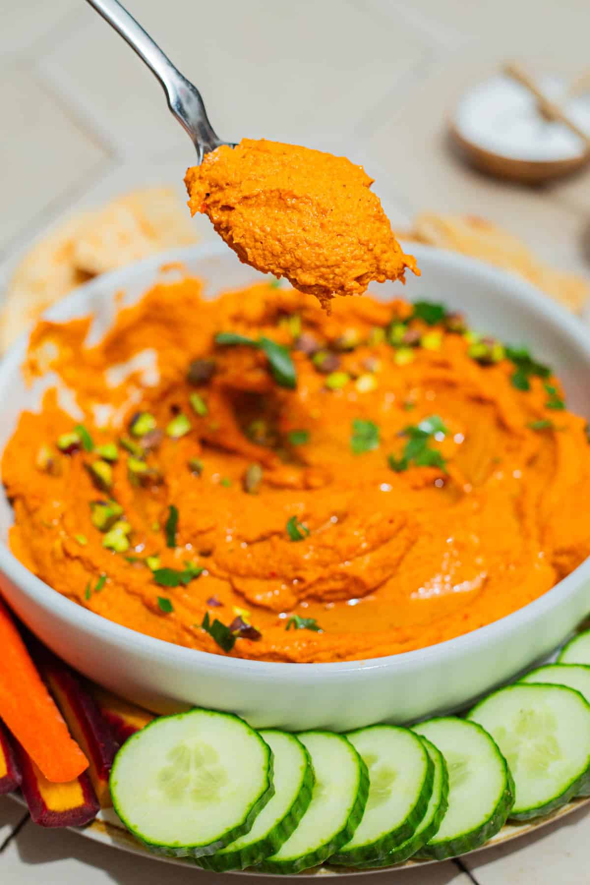 A close up photo of roasted carrot hummus on a spoon. In the background is the rest of the hummus in a bowl on a platter surrounded by pita bread triangles, carrot sticks and cucumber slices.