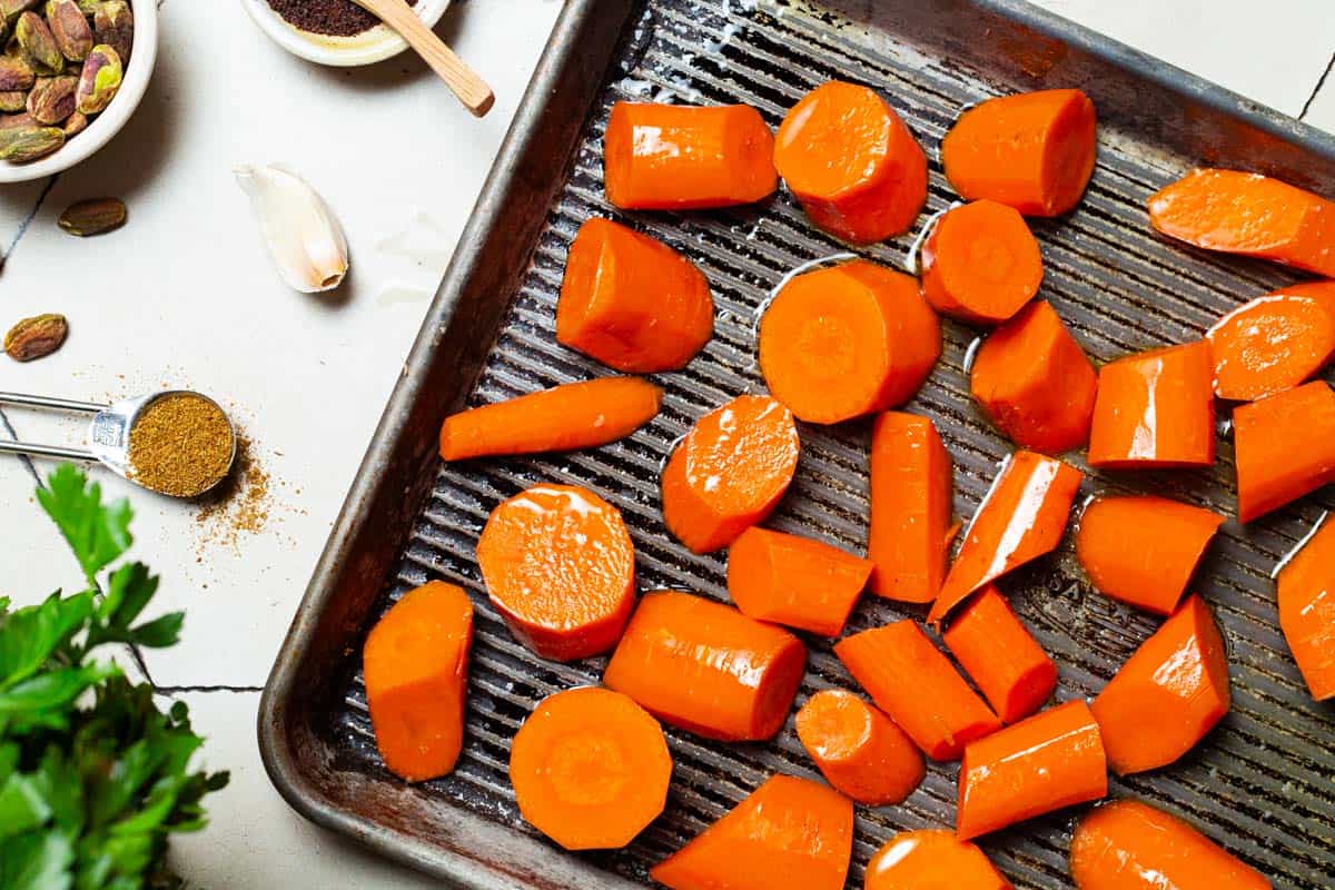 A close up of carrot pieces on a sheet pan surrounded by parsley, a measuring spoon with cumin, and bowls of pistachios and sumac.