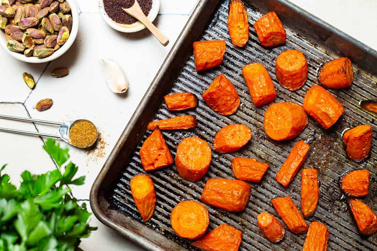 A close up of roasted carrot pieces on a sheet pan surrounded by parsley, a garlic clove, a measuring spoon with cumin, and bowls of pistachios and sumac.