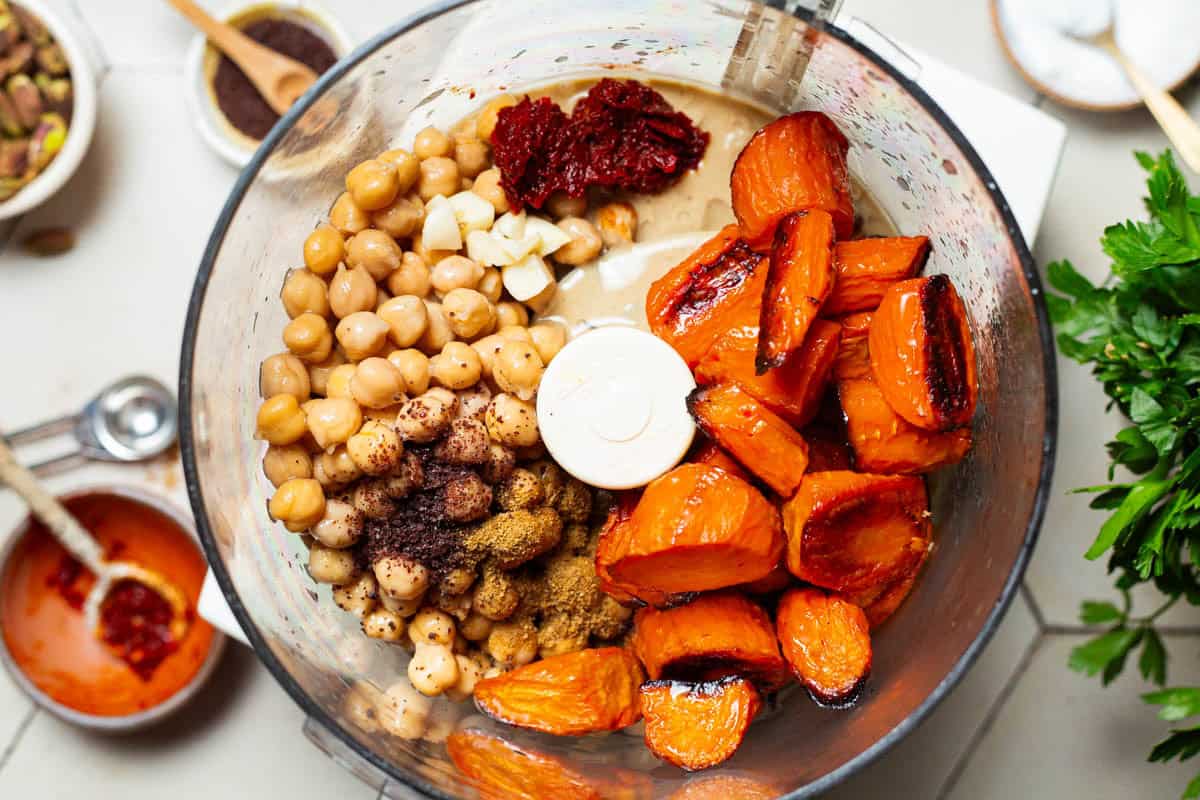 An overhead close up photo of the ingredients for the roasted carrot hummus in the bowl of a food processor just before being mixed together.