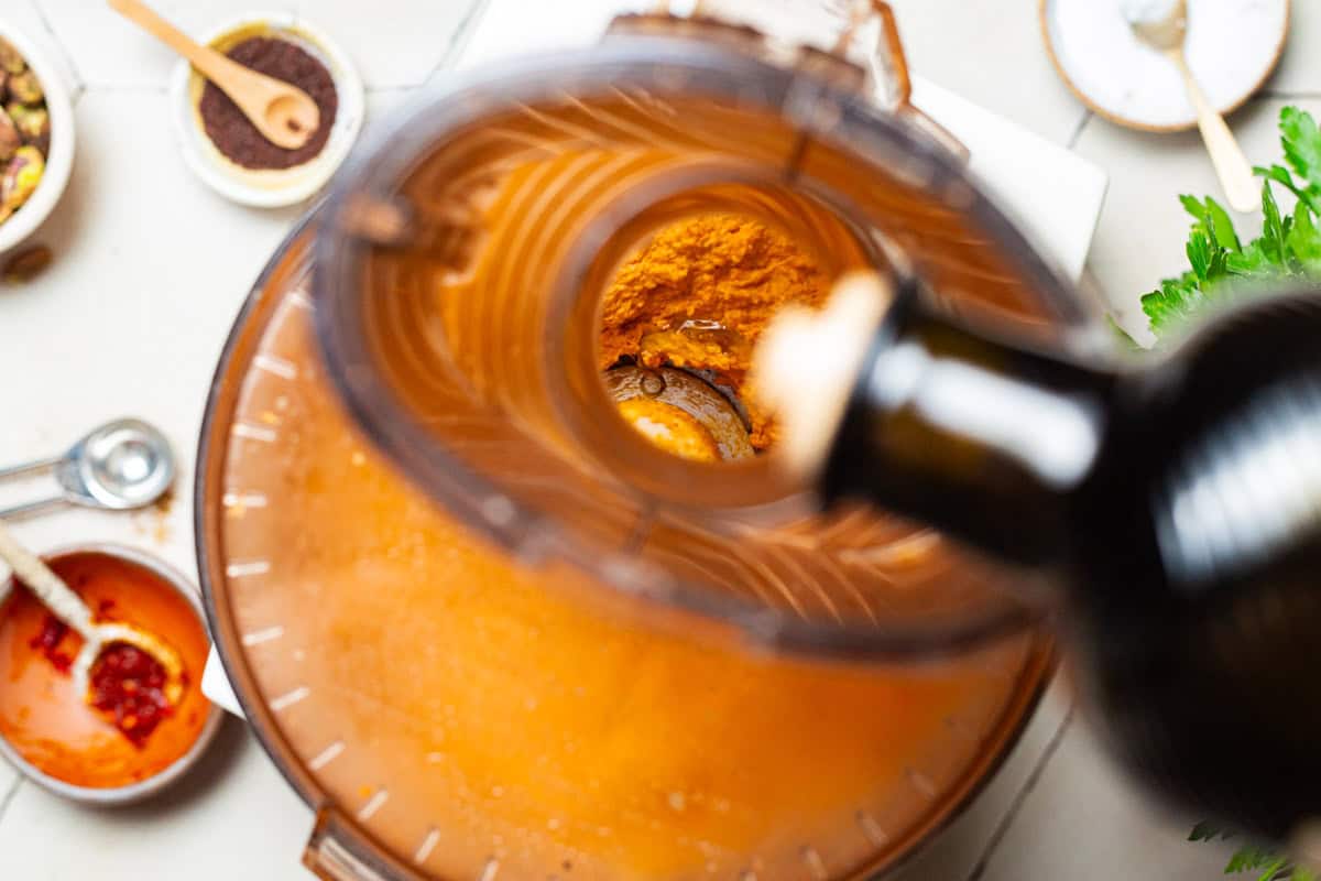 A close up of olive oil being poured from the bottle into the roasted carrot hummus in a food processor.