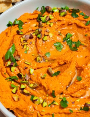 A close up of roasted carrot hummus topped with chopped pistachios and parsley in a bowl.