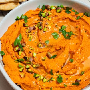 A close up of roasted carrot hummus topped with chopped pistachios and parsley in a bowl.