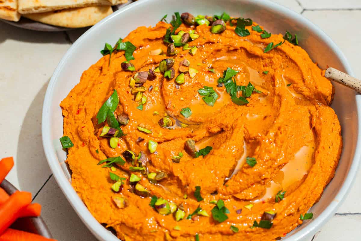 A close up of roasted carrot hummus topped with chopped pistachios and parsley in a bowl.