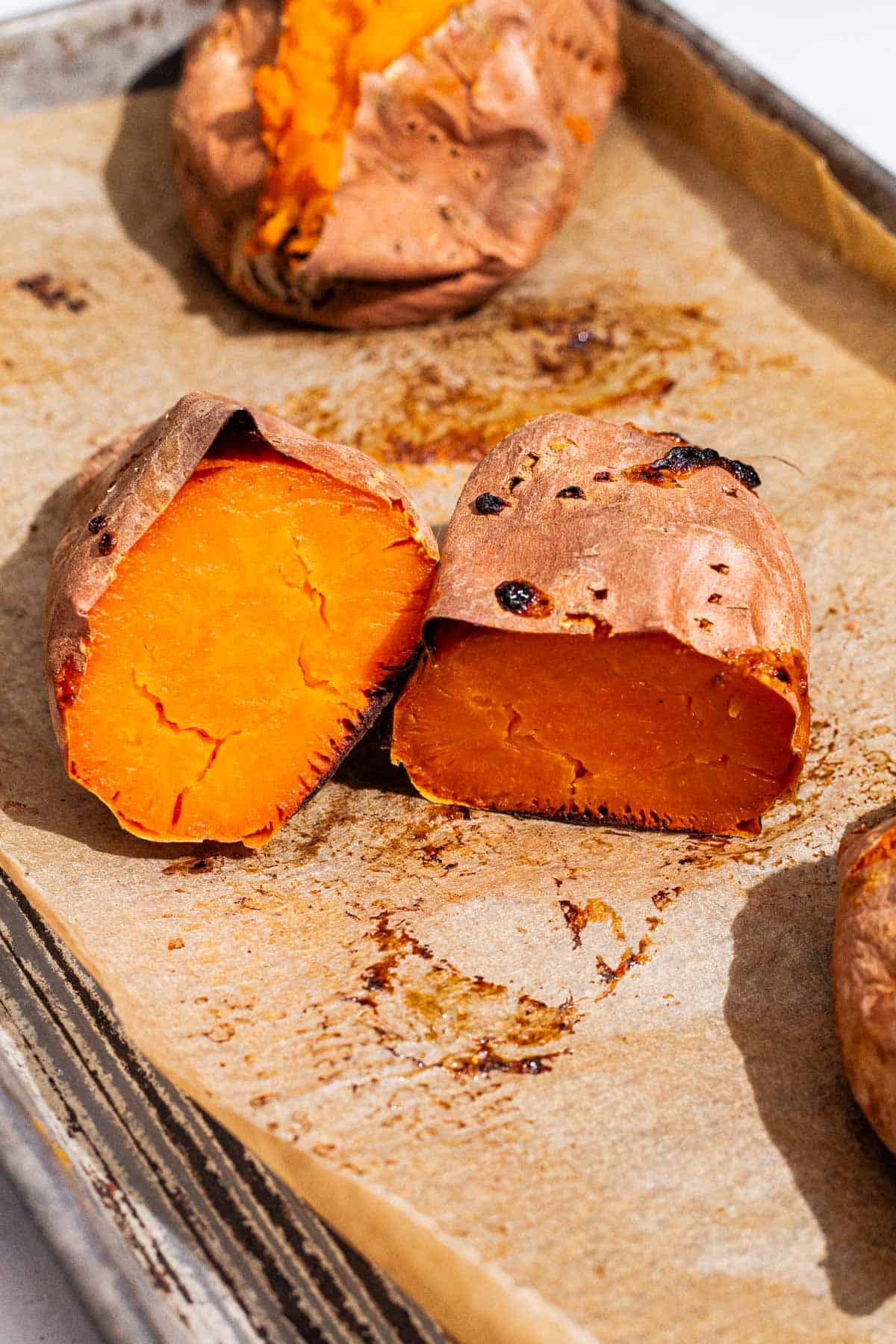 A close up of a roasted sweet potato cut in half on a parchment lined baking sheet, with another sweet potato in the background.