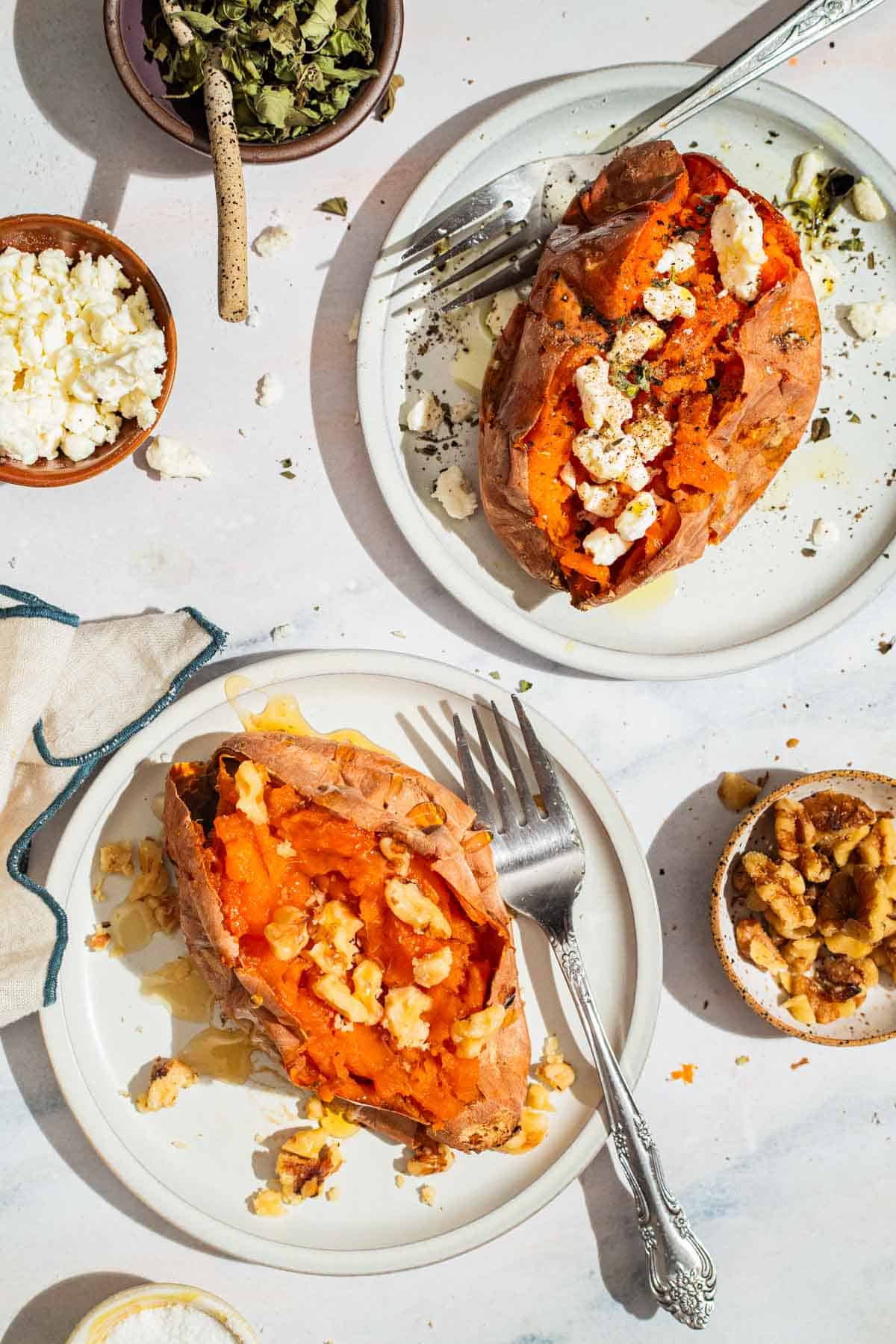 An overhead photo of one roasted sweet potato topped with honey and walnuts and one topped with spices and crumbled feta, each on a plate with a fork. Next to these are small bowls of crumbled feta, the spices, salt and walnuts.