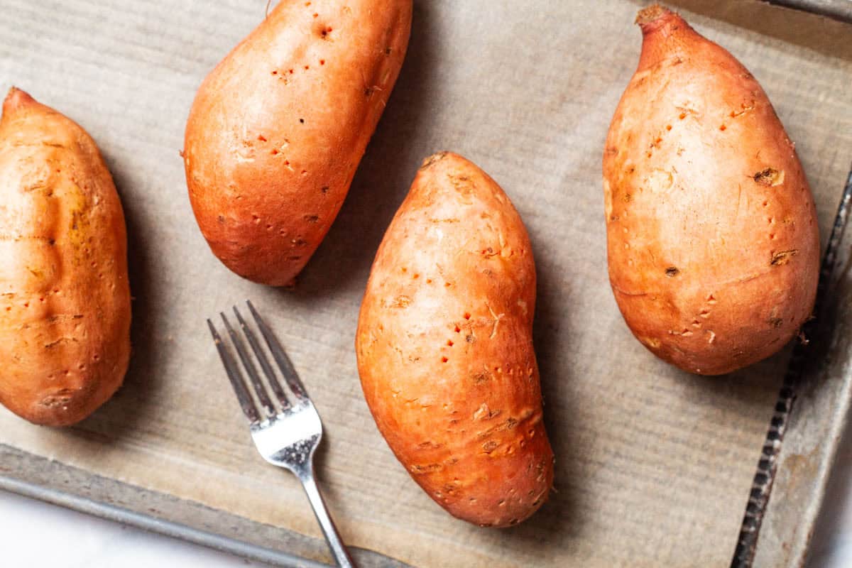 4 sweet potatoes on a parchment lined sheet pan just after being pierced with a fork.