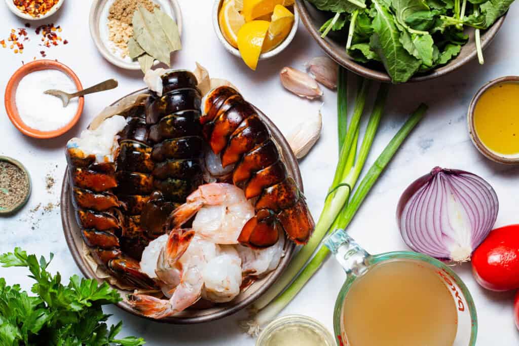 Ingredients for seafood stew including shrimp, lobster, lemons, bay leaves, ground ginger, garlic, salt, black pepper, olive oil, red onion, red pepper flakes, tomatoes, coriander, white wine, chicken broth, baby kale, green onions and parsley.