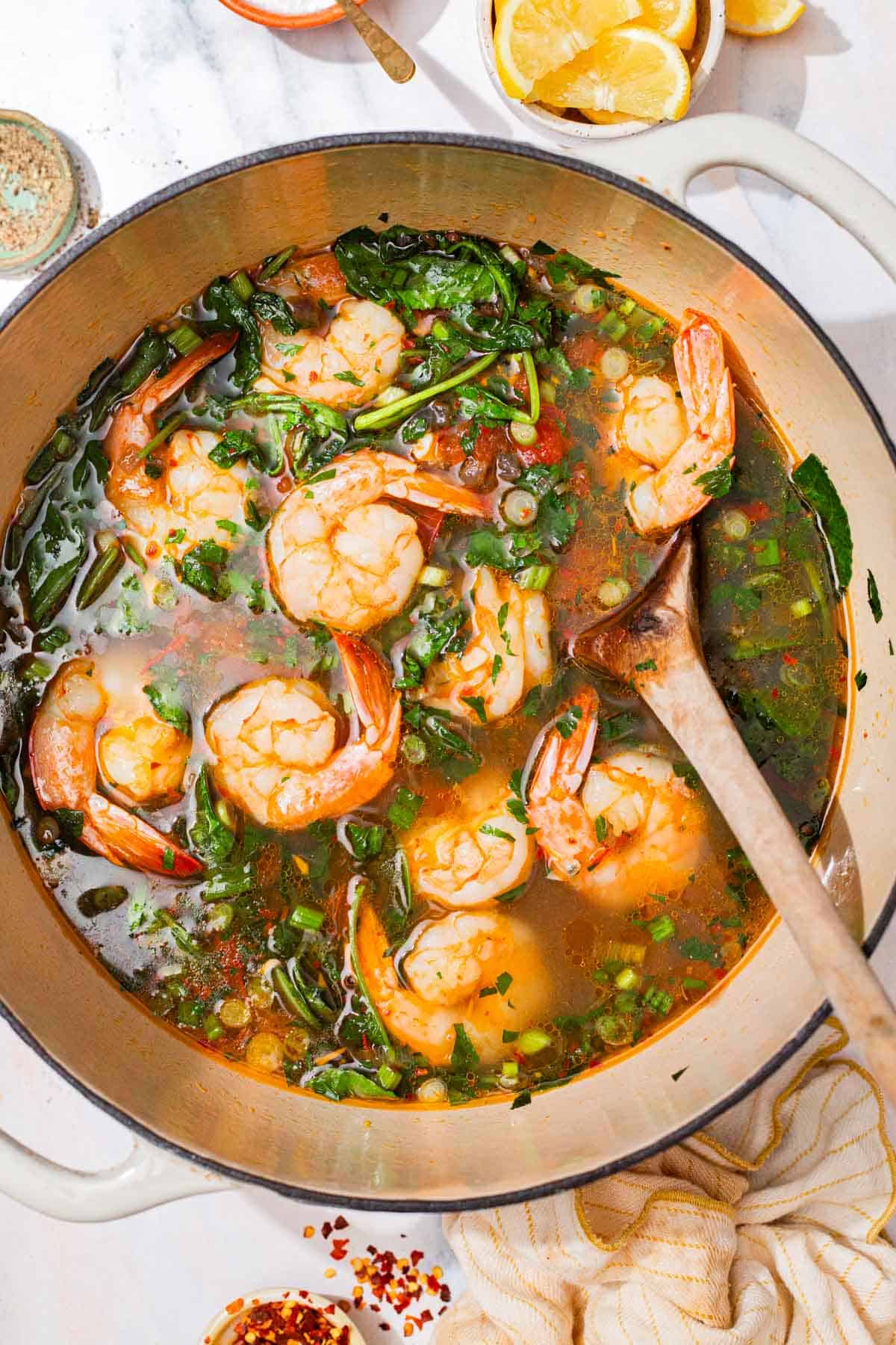 An overhead photo of seafood stew in a large pot with a wooden spoon. Next to this is a kitchen towel and small bowls of various other ingredients.