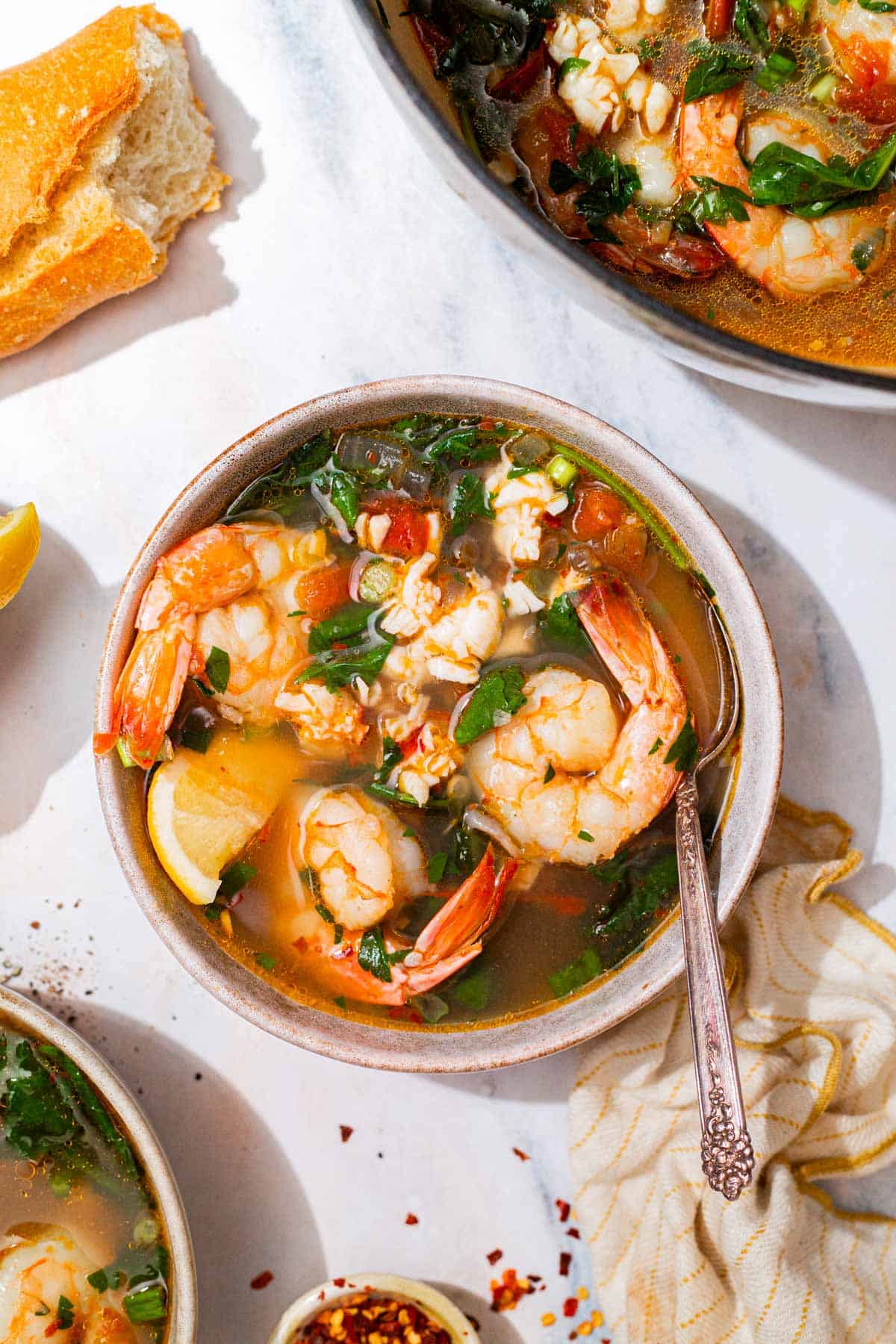 An overhead photo of a bowl of seafood stew with a spoon. Next to this is a kitchen towel, a piece of crusty bread, and the rest of the stew in a pot.