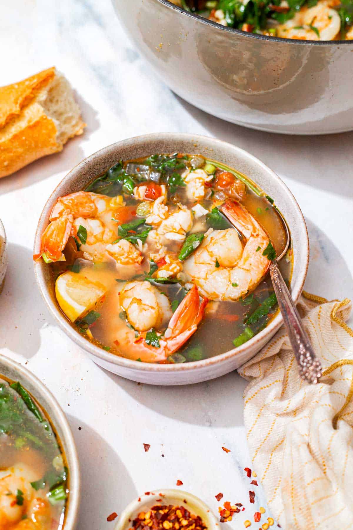 A close up of a bowl of seafood stew with a spoon. Next to this is a kitchen towel, a piece of crusty bread, a small bowl of red pepper flakes, and the rest of the stew in a pot.