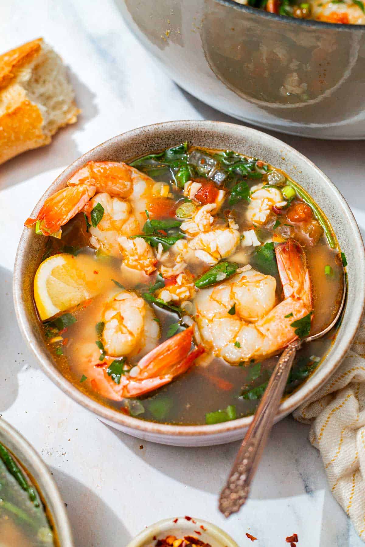 A close up of a bowl of seafood stew with a spoon. In the background is the pot with the rest of the stew, and a piece of crusty bread.